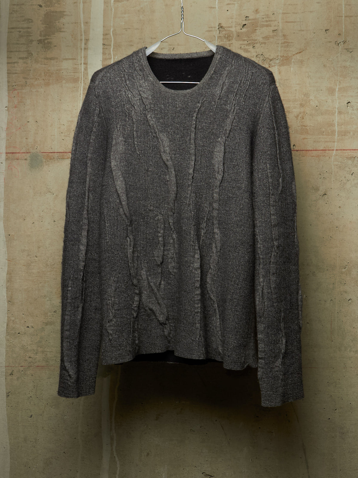 A dark gray textured knit sweater with vertical cable-like patterns, hanging on a metal hanger against a rustic concrete wall.