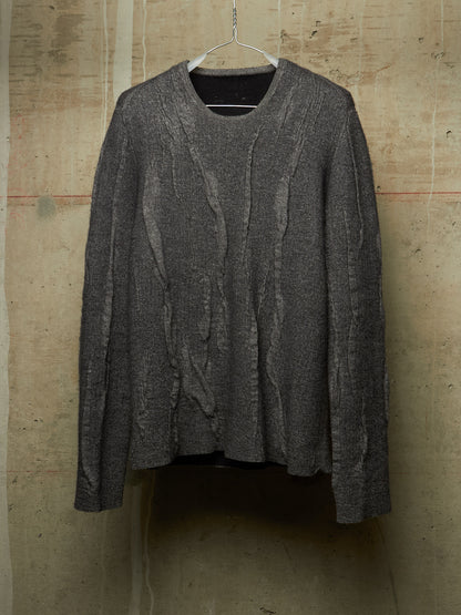 A dark gray textured knit sweater with vertical cable-like patterns, hanging on a metal hanger against a rustic concrete wall.