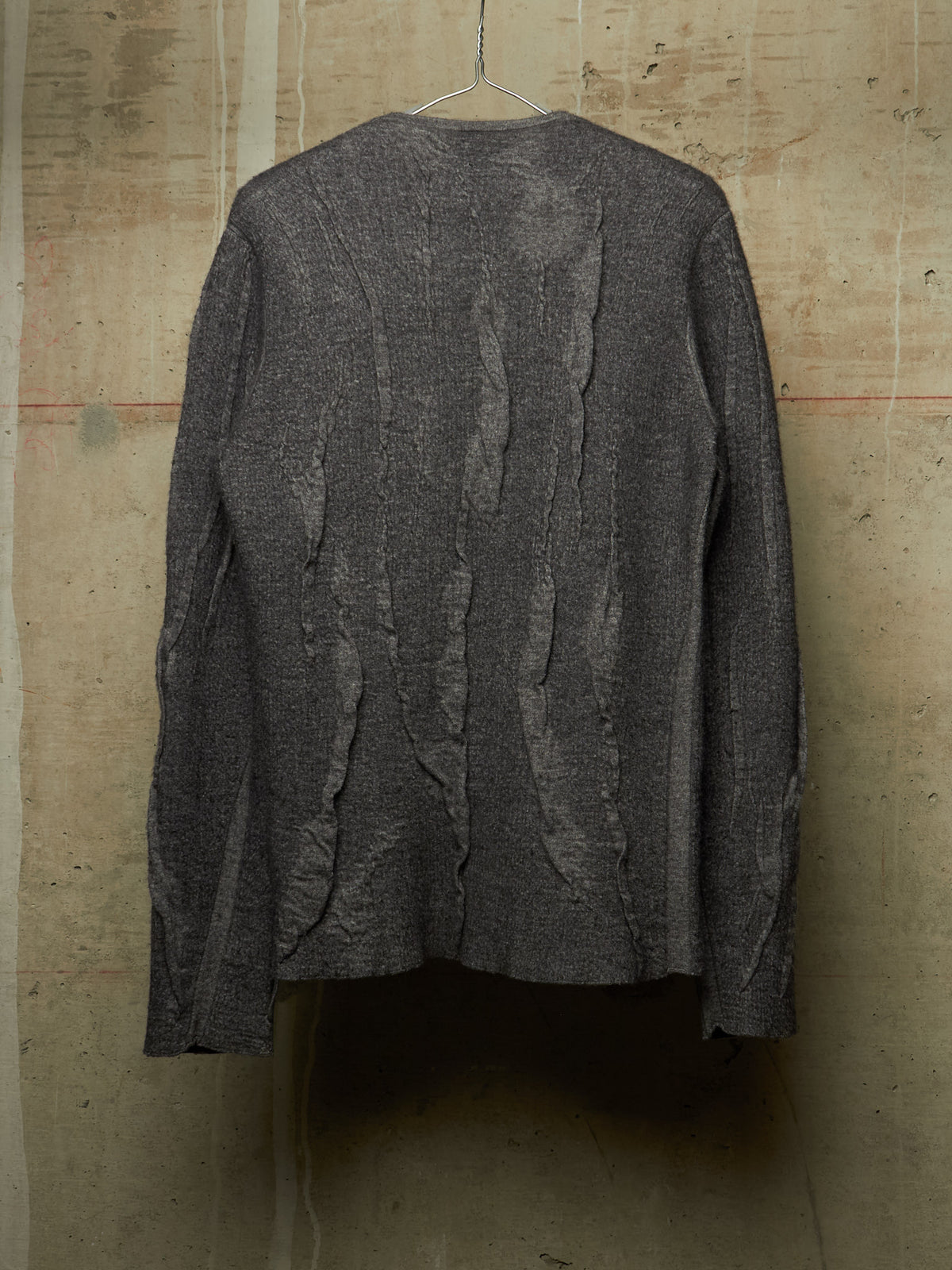 A dark gray textured knit cardigan sweater with vertical ruffled details, hanging on a wire hanger against a rustic concrete wall.