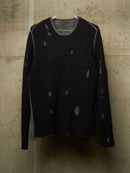 A black long-sleeve knit sweater with distressed, ripped details, hanging on a wire hanger against a textured concrete wall.
