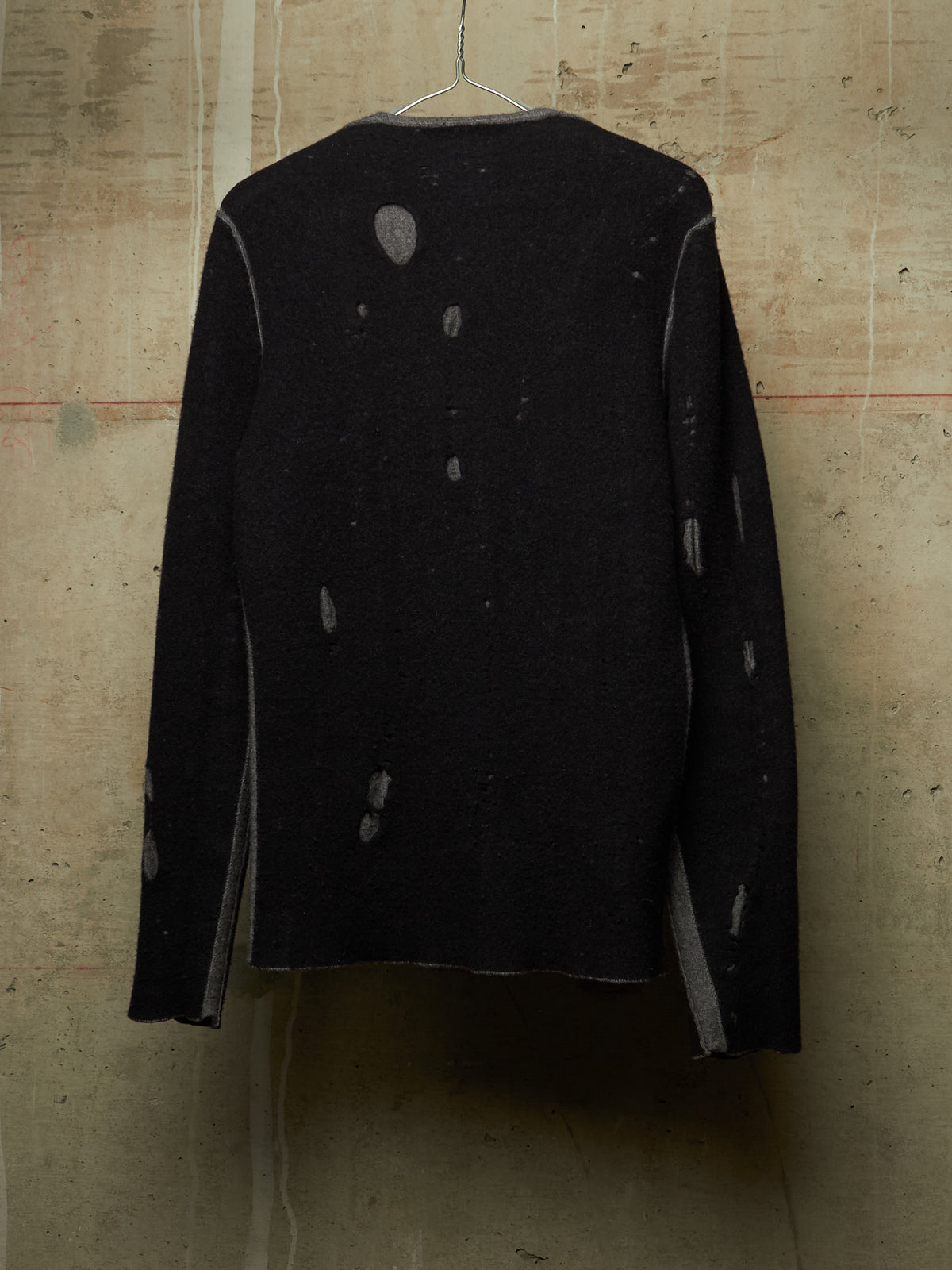 A black long-sleeve knit sweater with distressed, faded gray details, hanging on a wire hanger against a textured concrete wall.