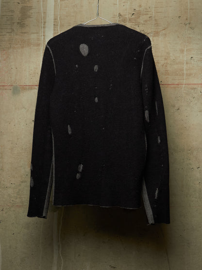 A black long-sleeve knit sweater with distressed, faded gray details, hanging on a wire hanger against a textured concrete wall.