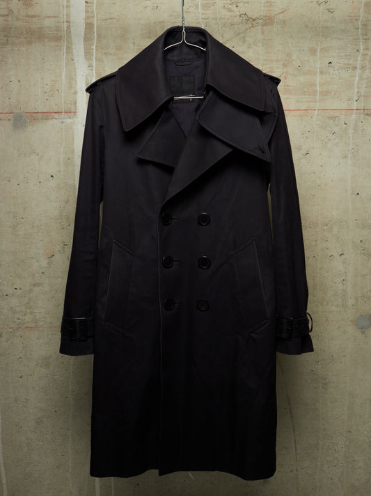 A black double-breasted trench coat with a wide collar and large front buttons, hanging on a wire hanger against a textured concrete wall.