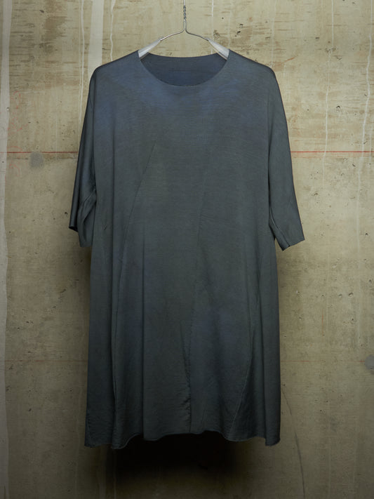 A loose-fitting, short-sleeved tunic dress in a dark charcoal gray color, hanging on a white wire hanger against a textured concrete wall.