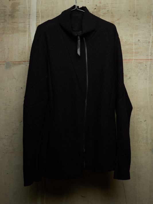 A black asymmetrical zip-up coat with a mock neck and long sleeves, hanging against a textured concrete wall.