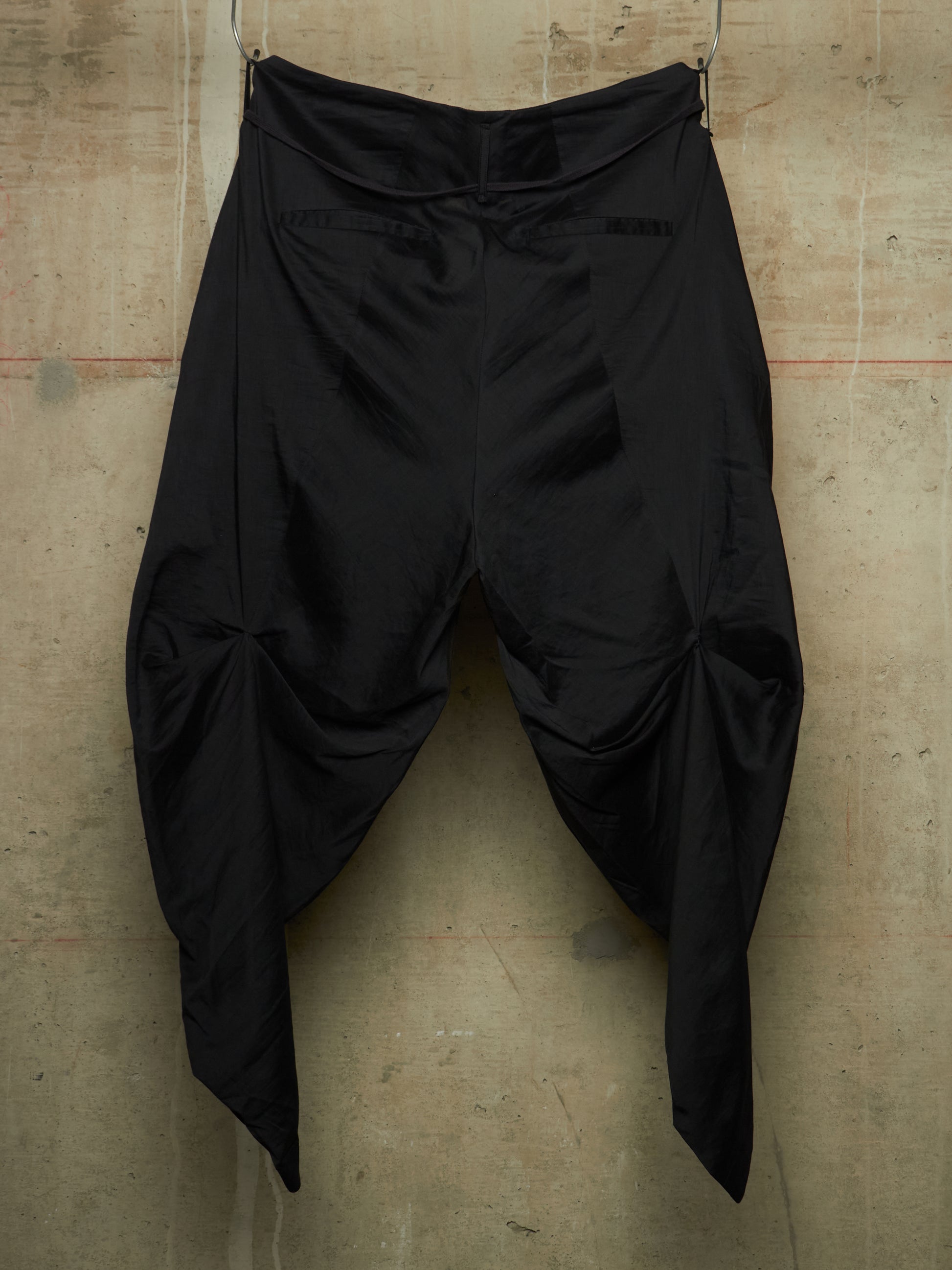 Black draped pants with a high waist and voluminous gathered legs, hanging against a textured concrete wall.