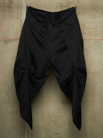 Black draped pants with a high waist and voluminous gathered legs, hanging against a textured concrete wall.
