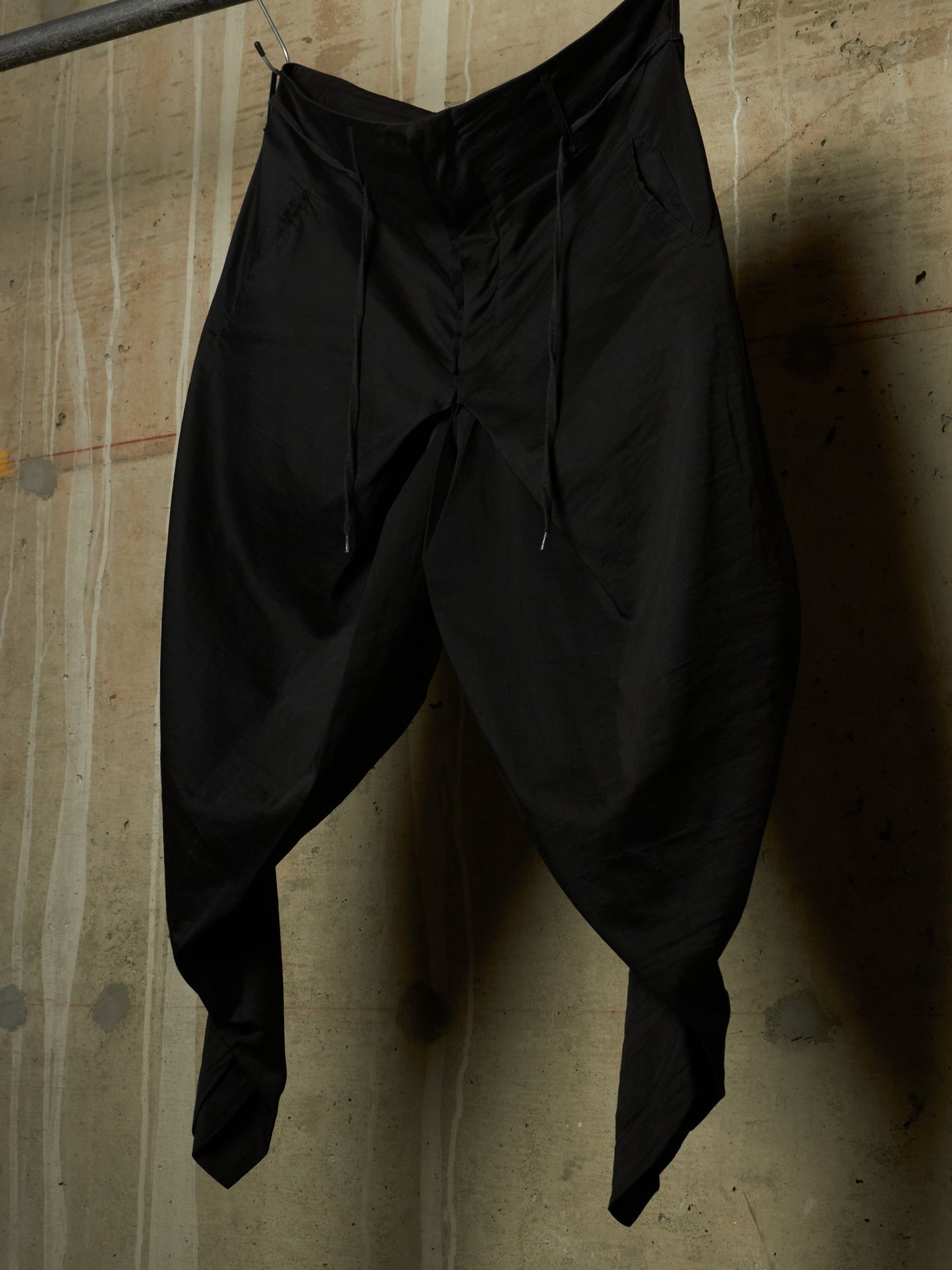 Black harem-style pants with a drawstring waist, hanging against a textured concrete wall.