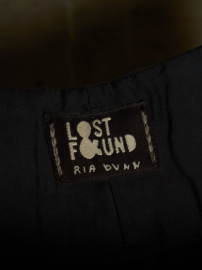 Close-up of a black fabric garment showing a sewn-in label that reads 'LOST FOUND' with a bowling pin icon and 'RIA DUNN' below.