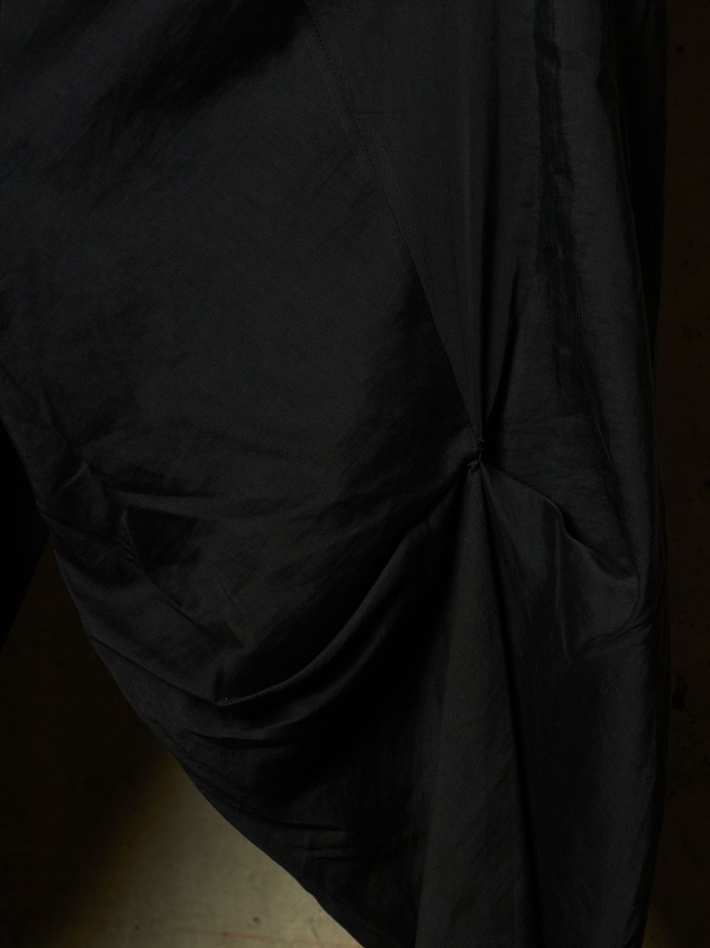 A close-up of draped black fabric with gathered folds and a gathered center detail, shown against a very dark background.