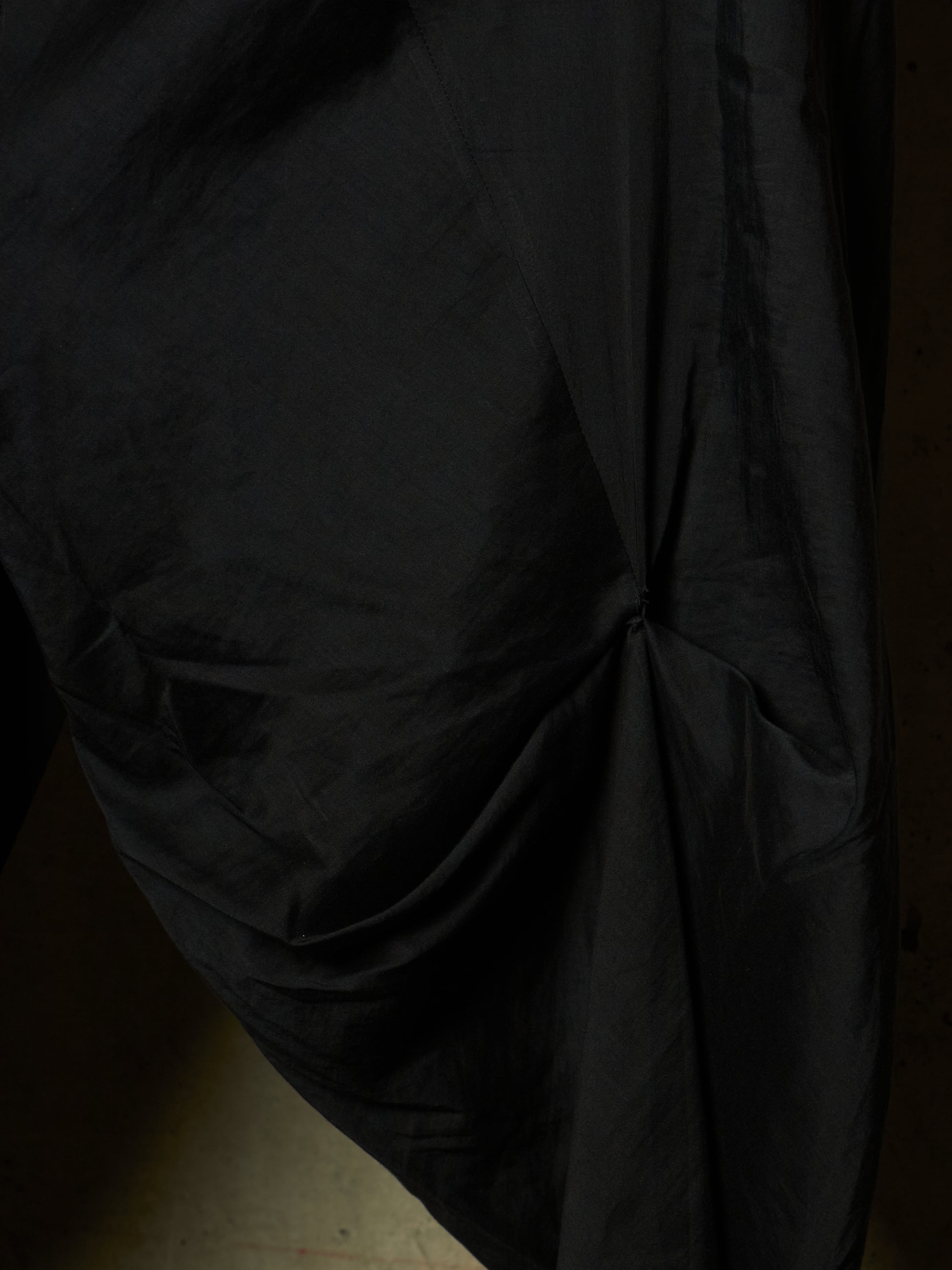 A close-up of draped black fabric with gathered folds and a gathered center detail, shown against a very dark background.