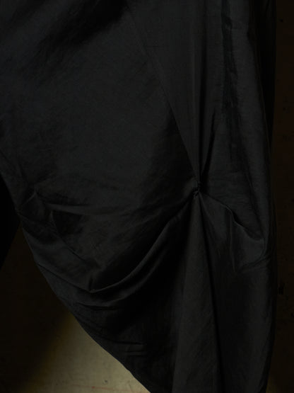 A close-up of draped black fabric with gathered folds and a gathered center detail, shown against a very dark background.