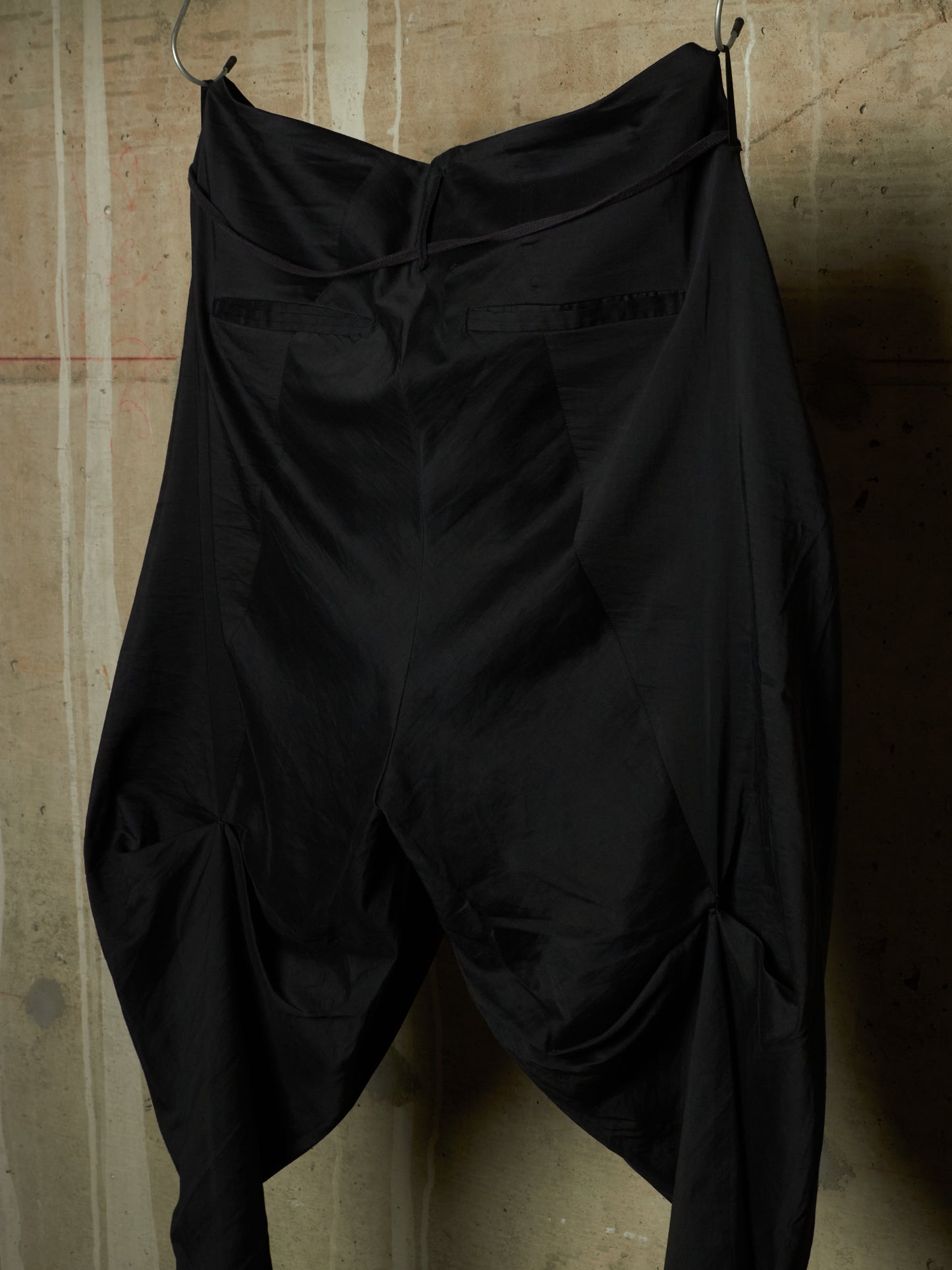 Black draped pants with a high gathered waist and side slit details, hanging against a textured concrete wall.