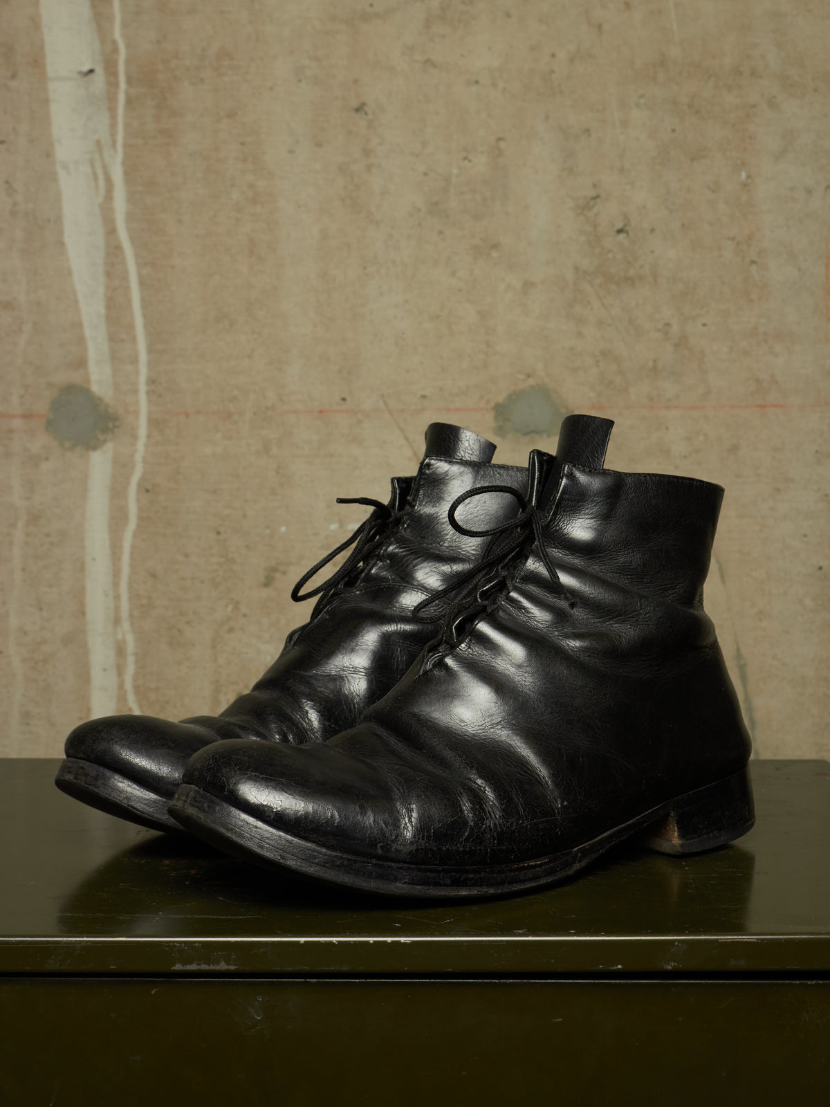 A pair of black leather lace-up ankle boots with a slightly worn, vintage aesthetic, resting on a dark green surface against a textured beige wall.