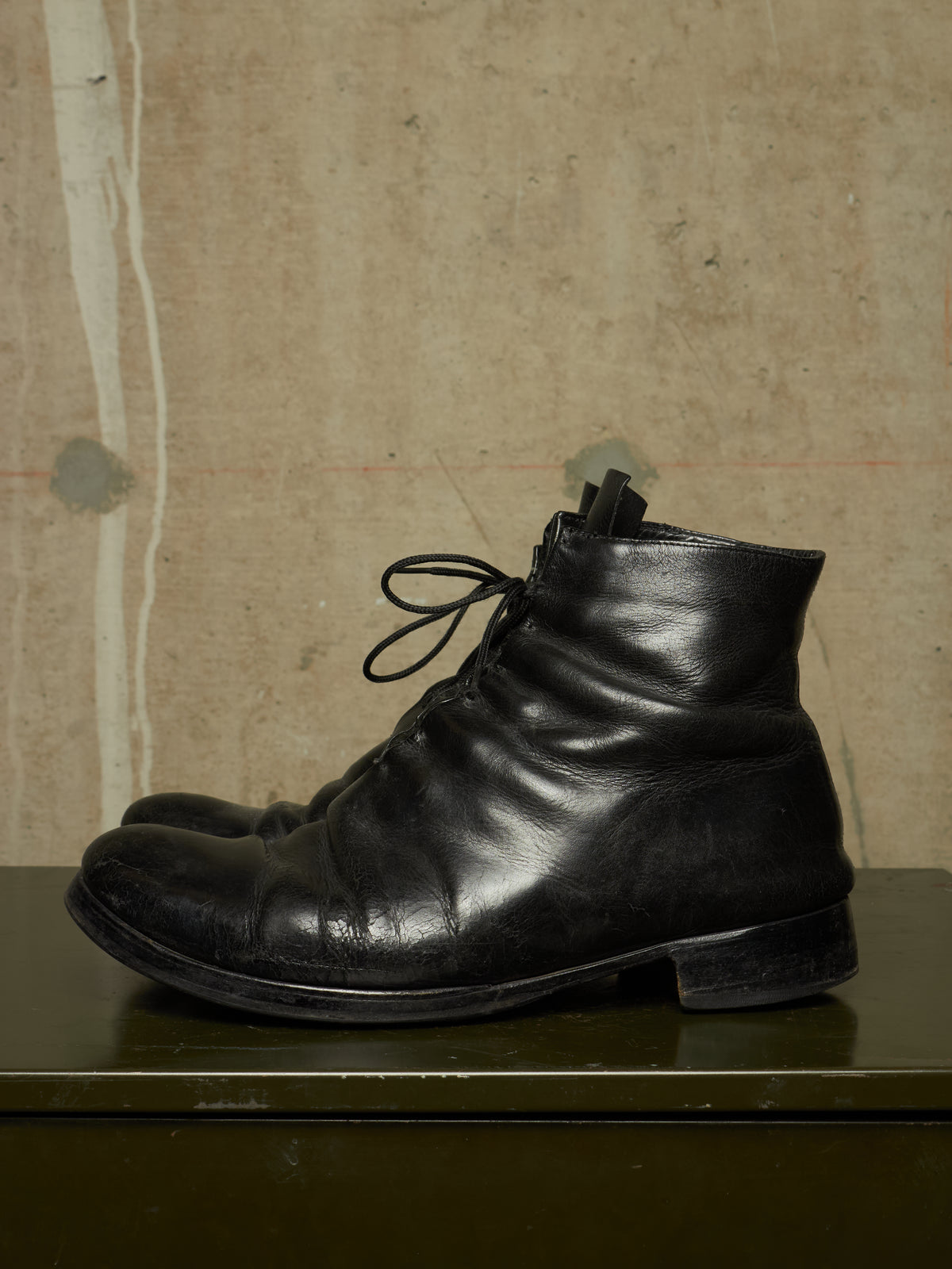 A pair of black leather lace-up ankle boots with a distressed, scuffed finish, displayed on a dark green surface against a textured beige concrete wall.