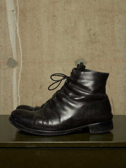 A pair of black leather lace-up ankle boots with a distressed, scuffed finish, displayed on a dark green surface against a textured beige concrete wall.