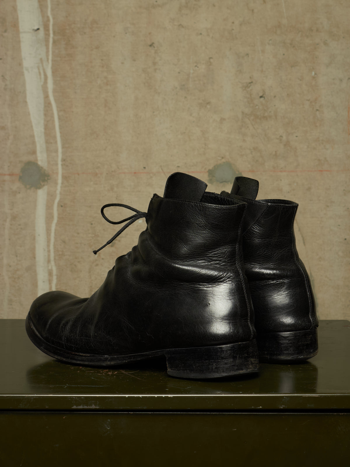 A pair of black leather ankle boots with a low block heel and lace-up front, displayed on a dark green surface against a textured beige wall.