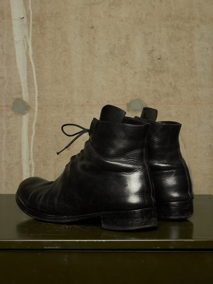 A pair of black leather ankle boots with a low block heel and lace-up front, displayed on a dark green surface against a textured beige wall.