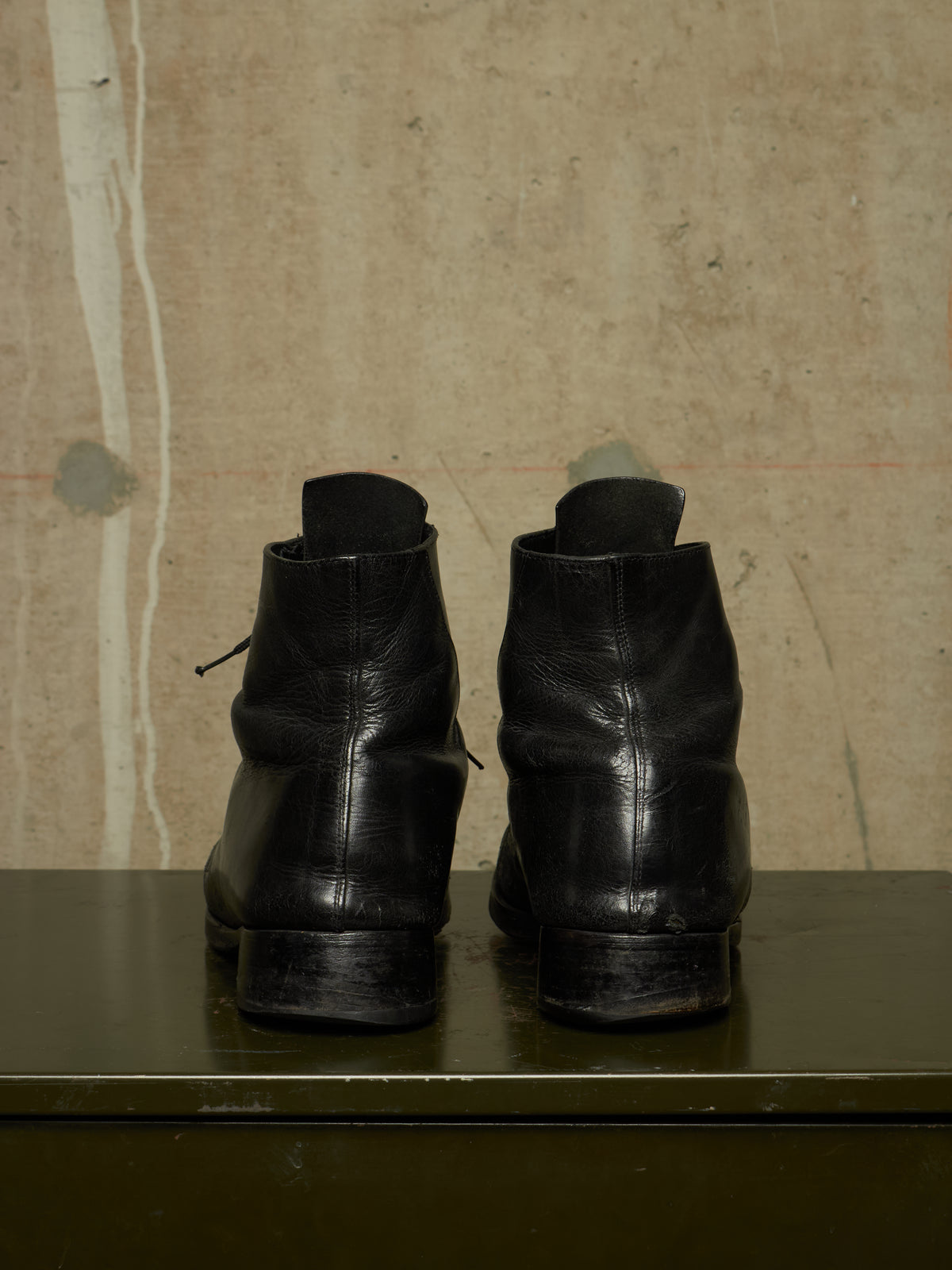 A pair of black leather ankle boots shown from the back, resting on a dark green surface against a textured beige wall.