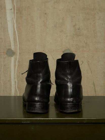 A pair of black leather ankle boots shown from the back, resting on a dark green surface against a textured beige wall.