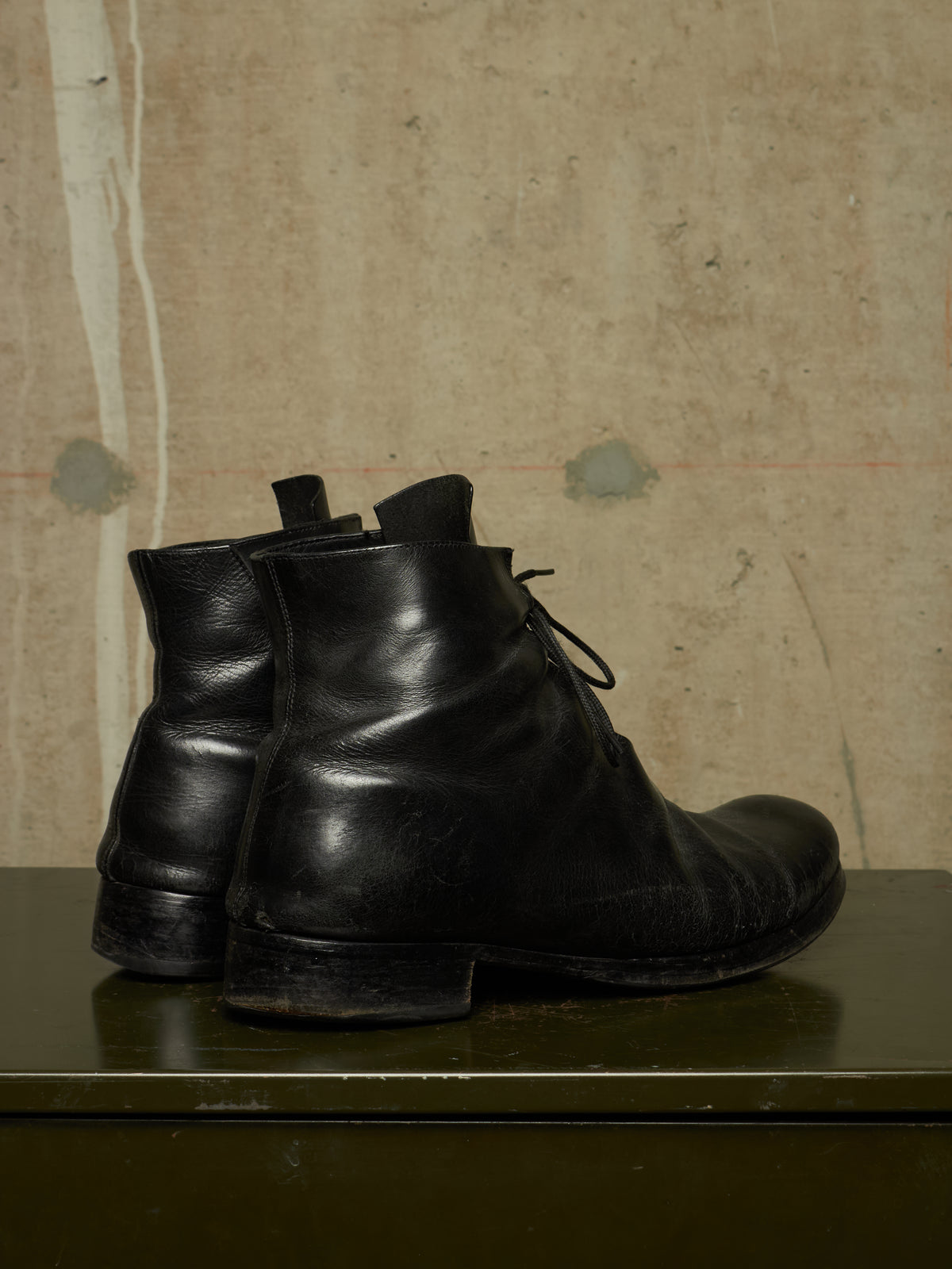 A pair of black leather lace-up ankle boots standing on a dark green surface against a textured beige wall.