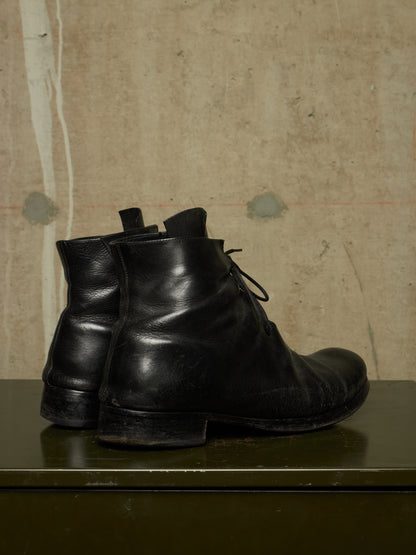 A pair of black leather lace-up ankle boots standing on a dark green surface against a textured beige wall.