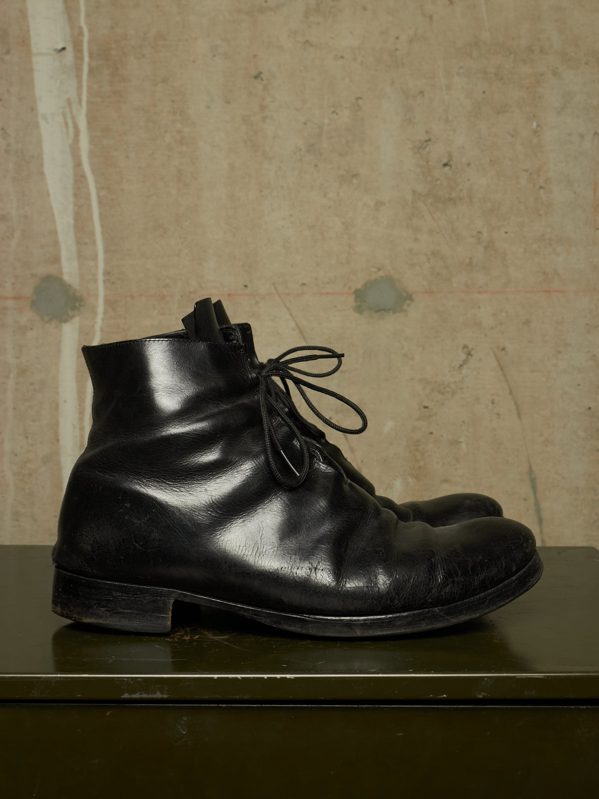 A pair of black leather lace-up ankle boots resting on a dark green surface against a textured beige wall.
