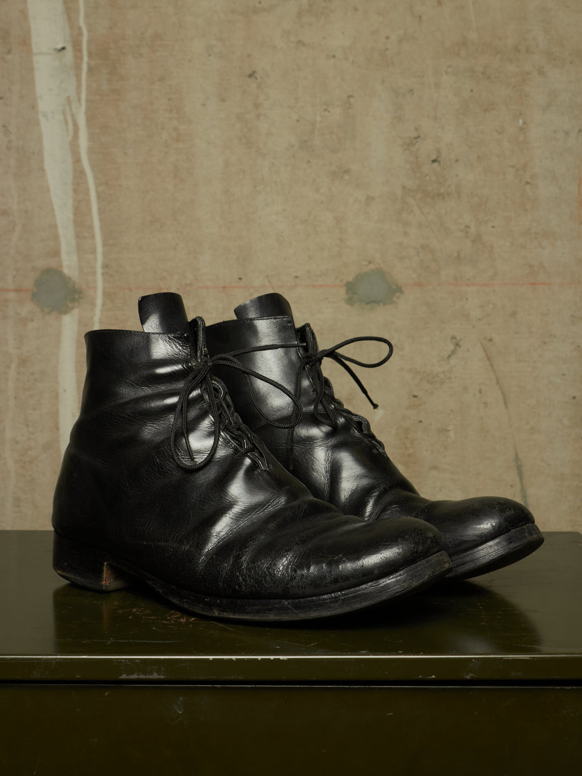 A pair of black leather lace-up ankle boots with a distressed, worn finish, resting on a dark green surface against a textured beige background.