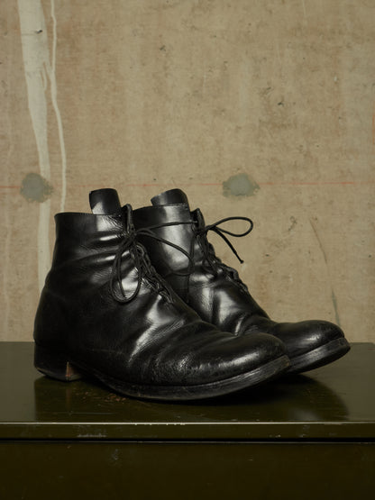 A pair of black leather lace-up ankle boots with a distressed, worn finish, resting on a dark green surface against a textured beige background.