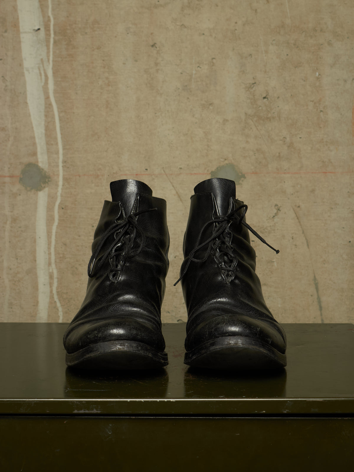 A pair of black leather lace-up ankle boots with a distressed, worn look, displayed on a dark green surface against a textured beige wall.