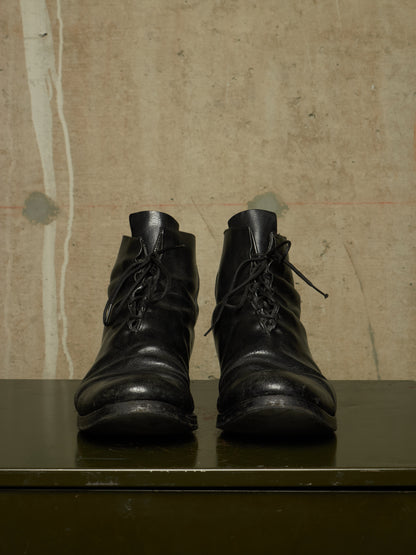 A pair of black leather lace-up ankle boots with a distressed, worn look, displayed on a dark green surface against a textured beige wall.