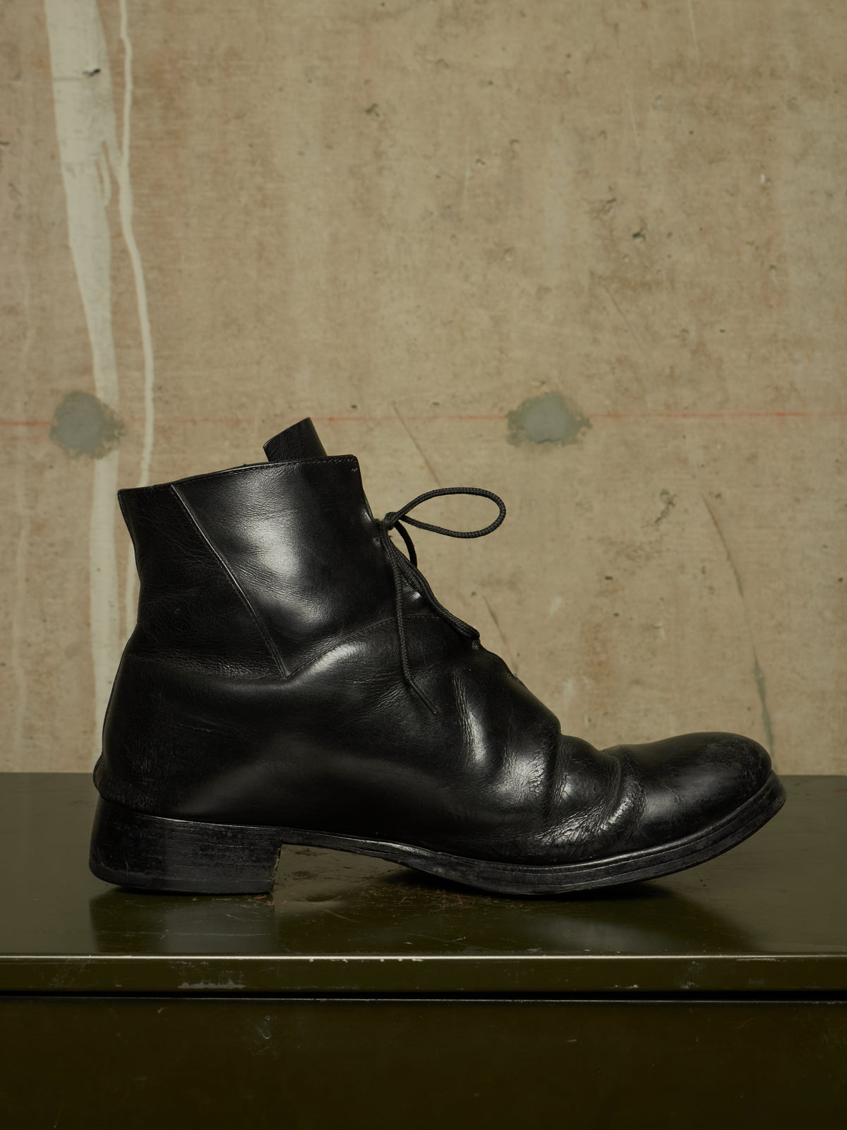 A single black leather lace-up ankle boot with a low block heel, shown in side profile on a dark green surface against a textured beige wall.