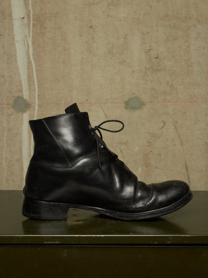 A single black leather lace-up ankle boot with a low block heel, shown in side profile on a dark green surface against a textured beige wall.