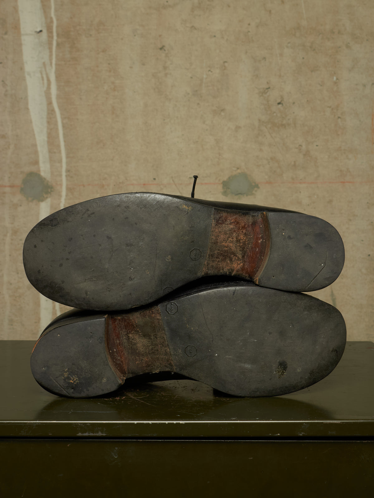 The soles of a pair of dark-colored shoes stacked on top of each other, showing a worn brown heel strip and dark rubber outsoles, set against a textured beige wall.