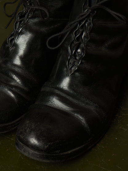 A close-up shot of a pair of black leather lace-up boots with a glossy finish, resting on a dark olive green textured surface.