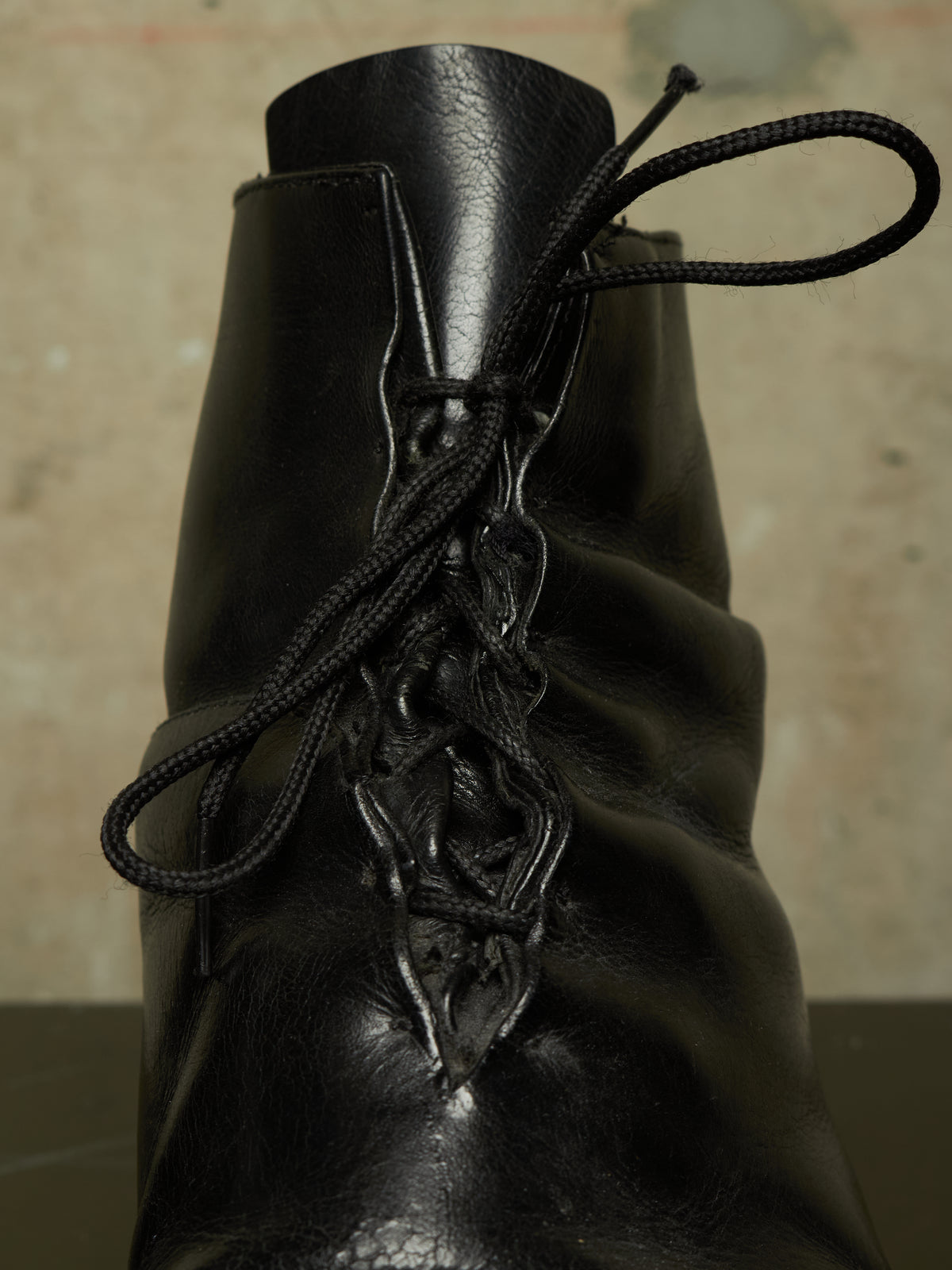 A close-up of a black leather lace-up ankle boot, showing the texture of the leather and the black laces threaded through decorative eyelets, set against a neutral, out-of-focus background.