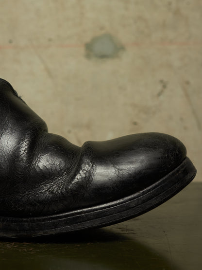 A close-up of a black leather boot toe showing the textured leather grain and thick black sole, set against a neutral concrete background.