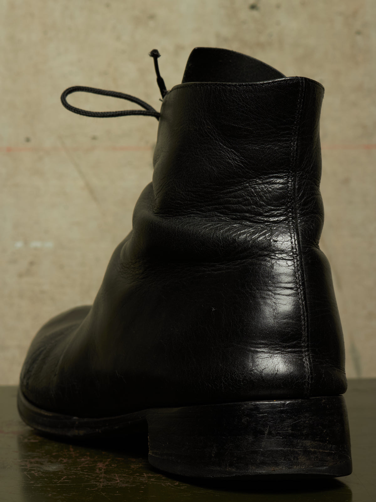 A close-up shot of a black leather ankle boot from the back, showing the heel, sole, and laces, set against a textured beige background.