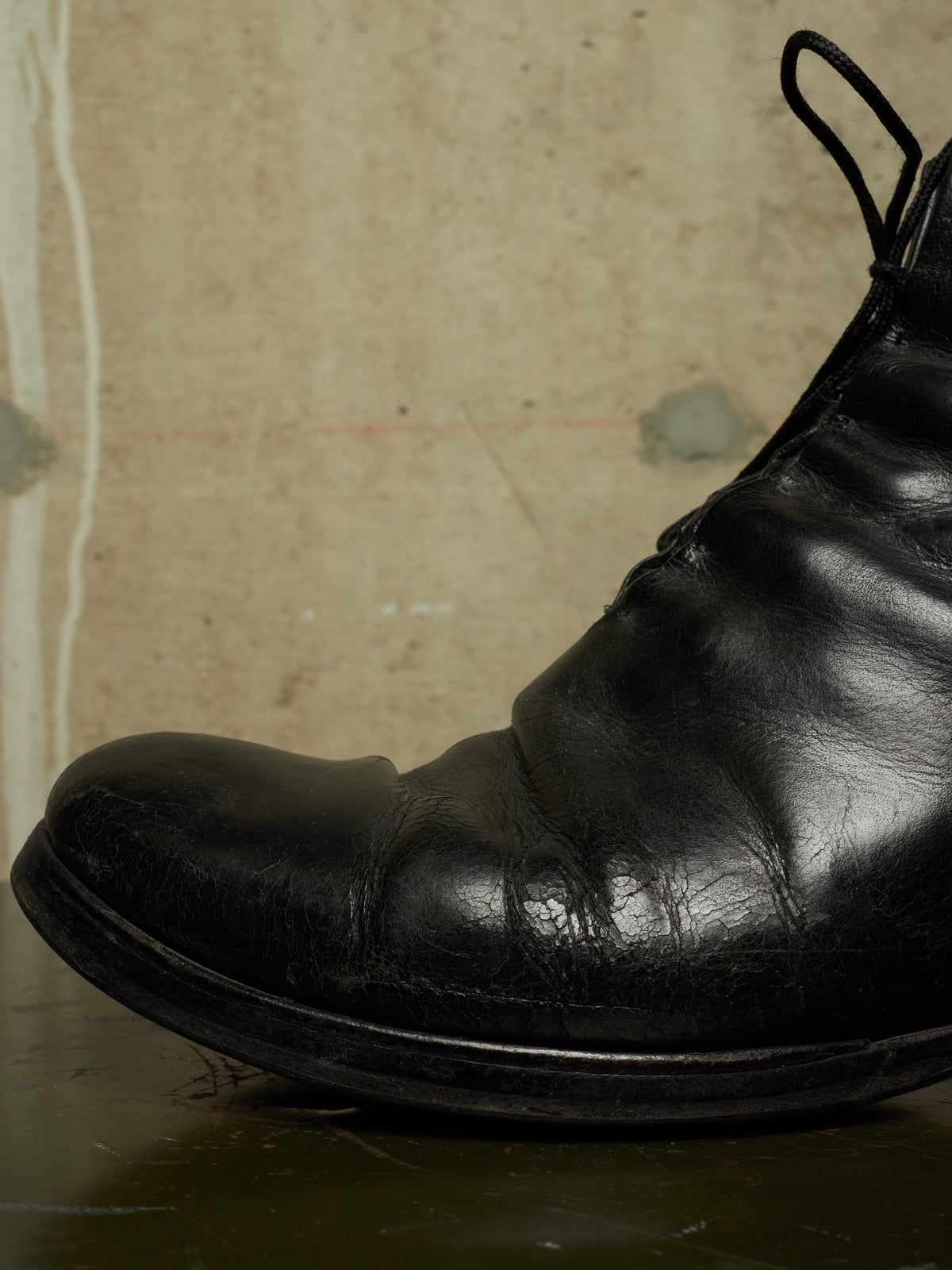A close-up of a black leather ankle boot with a glossy finish, showing the toe and side profile against a textured beige background.