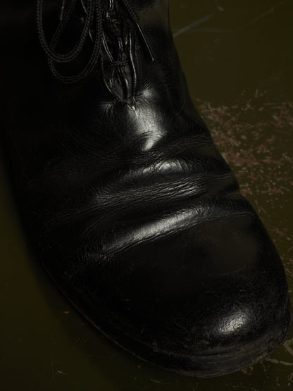 A close-up of a black leather lace-up boot or shoe with a glossy finish, showing the toe and side against a dark olive-green background.