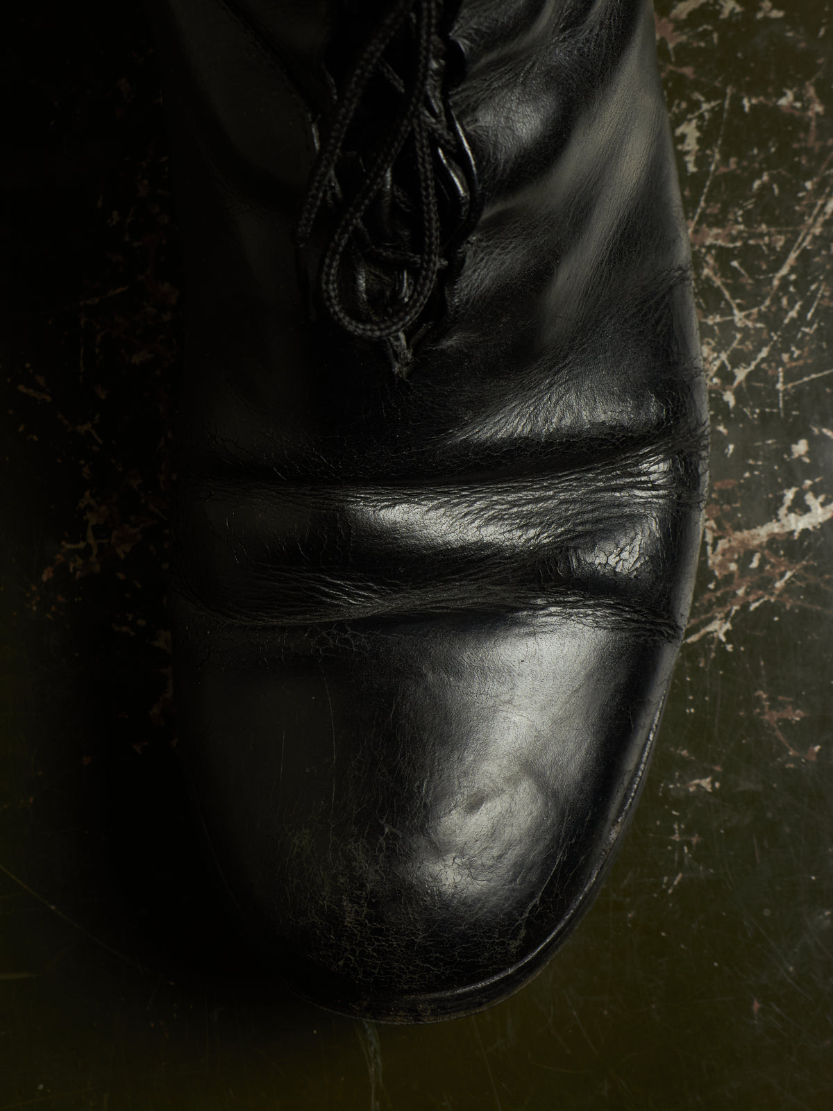 A close-up of a black leather lace-up boot resting on a dark, textured surface.
