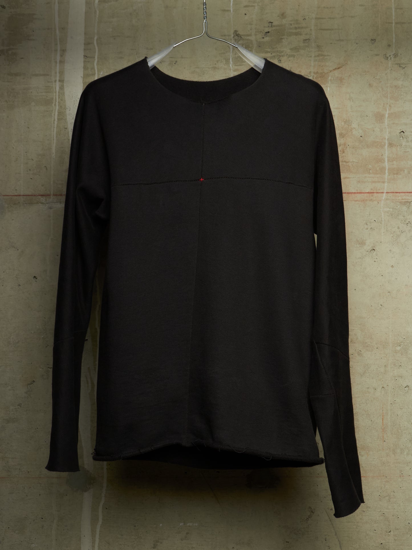 A black long-sleeve top hanging on a metal hanger against a textured concrete wall. The top features a subtle horizontal seam across the chest and a tiny red accent dot near the center.
