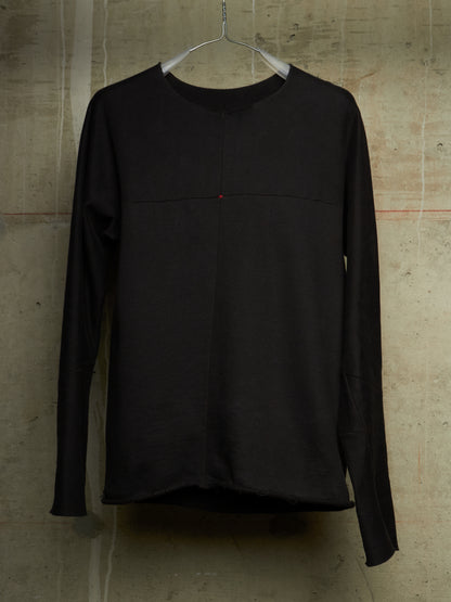 A black long-sleeve top hanging on a metal hanger against a textured concrete wall. The top features a subtle horizontal seam across the chest and a tiny red accent dot near the center.
