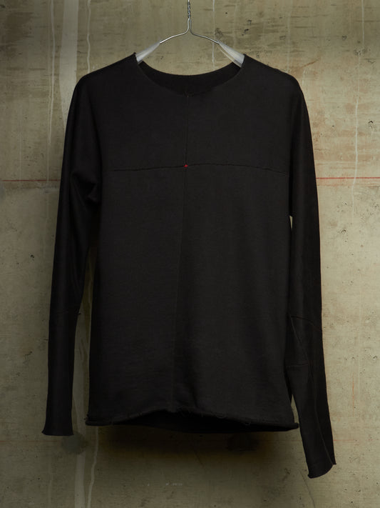A black long-sleeve top hanging on a metal hanger against a textured concrete wall. The top features a subtle horizontal seam across the chest and a tiny red accent dot near the center.