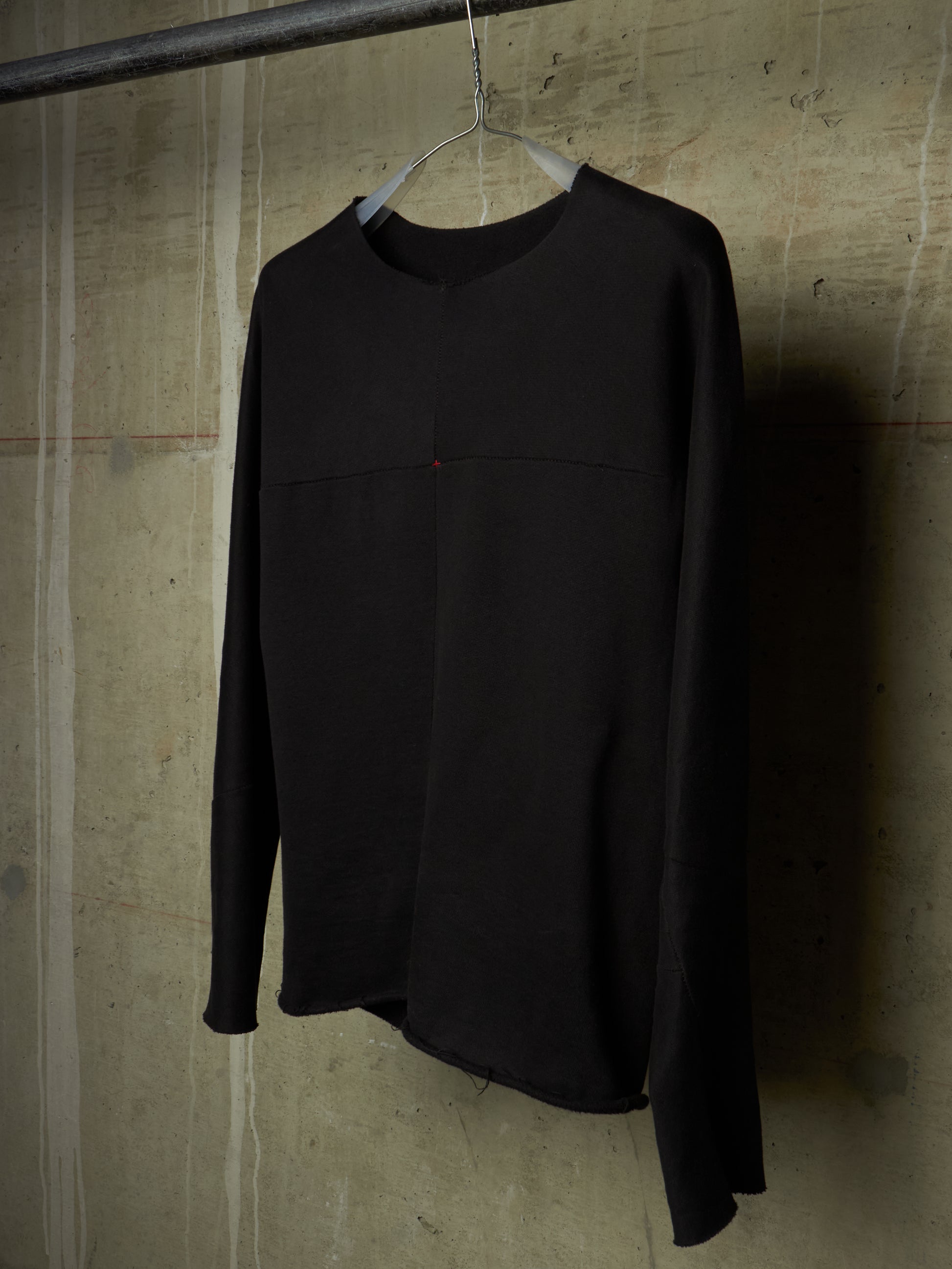A black long-sleeve top hanging on a wire hanger from a metal rod, set against a textured concrete wall.
