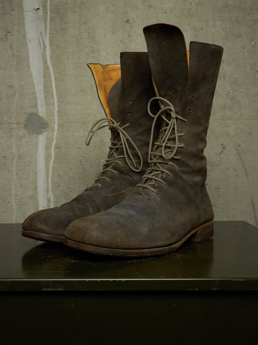 A pair of tall, dark brown distressed leather lace-up boots with tan interiors, displayed on a dark surface against a textured concrete wall.