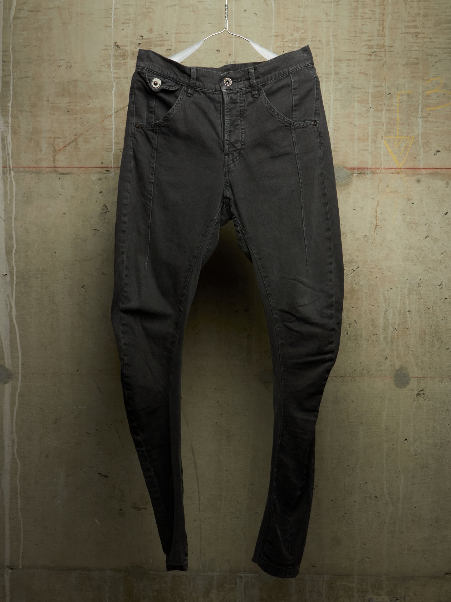 A pair of slim-fit black denim jeans hanging from a white plastic hanger against a textured concrete wall.
