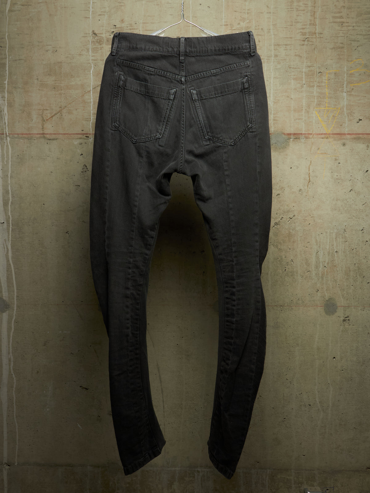A pair of faded black denim jeans hanging from a wire hanger against a textured concrete wall, showing the back pockets and leg seams.