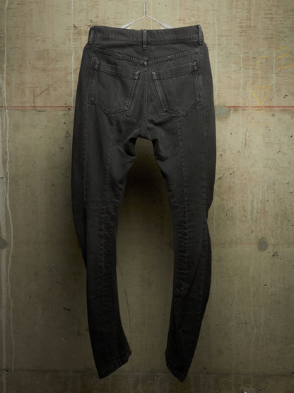 A pair of faded black denim jeans hanging from a wire hanger against a textured concrete wall, showing the back pockets and leg seams.