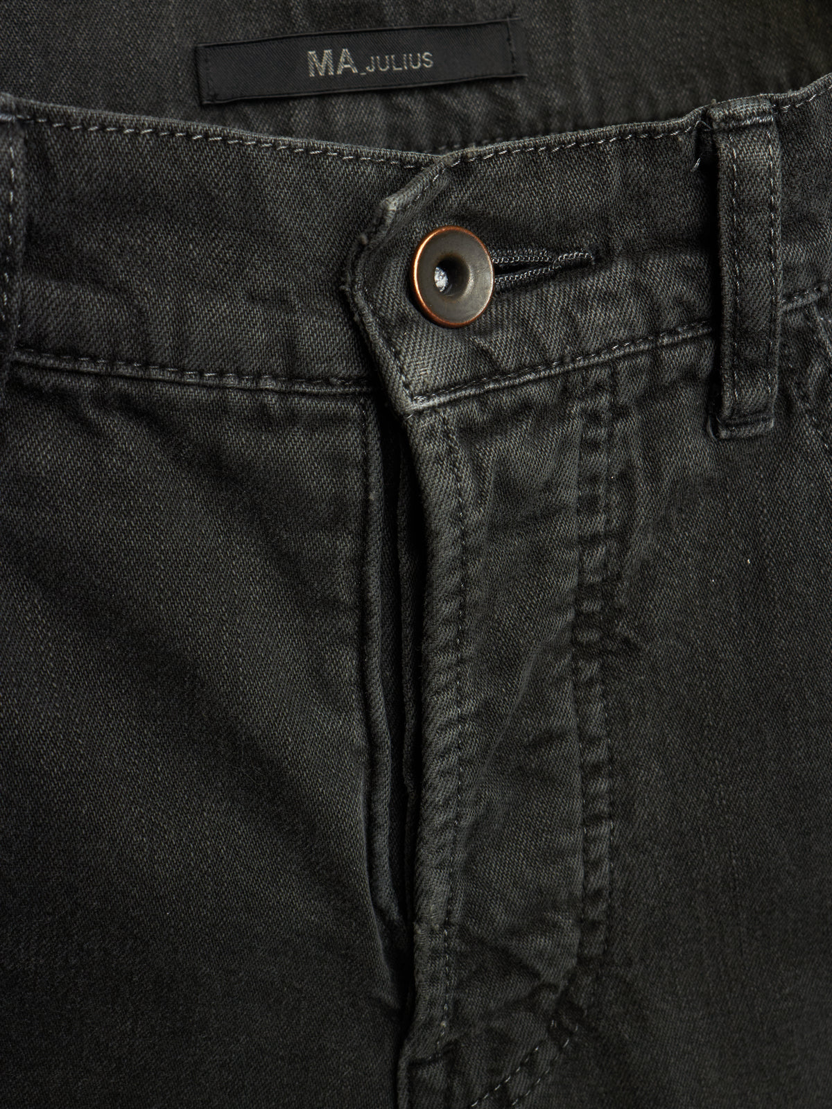 Close-up of black denim jeans showing the button closure, belt loop, and waistband label with the brand name MA JULIUS.